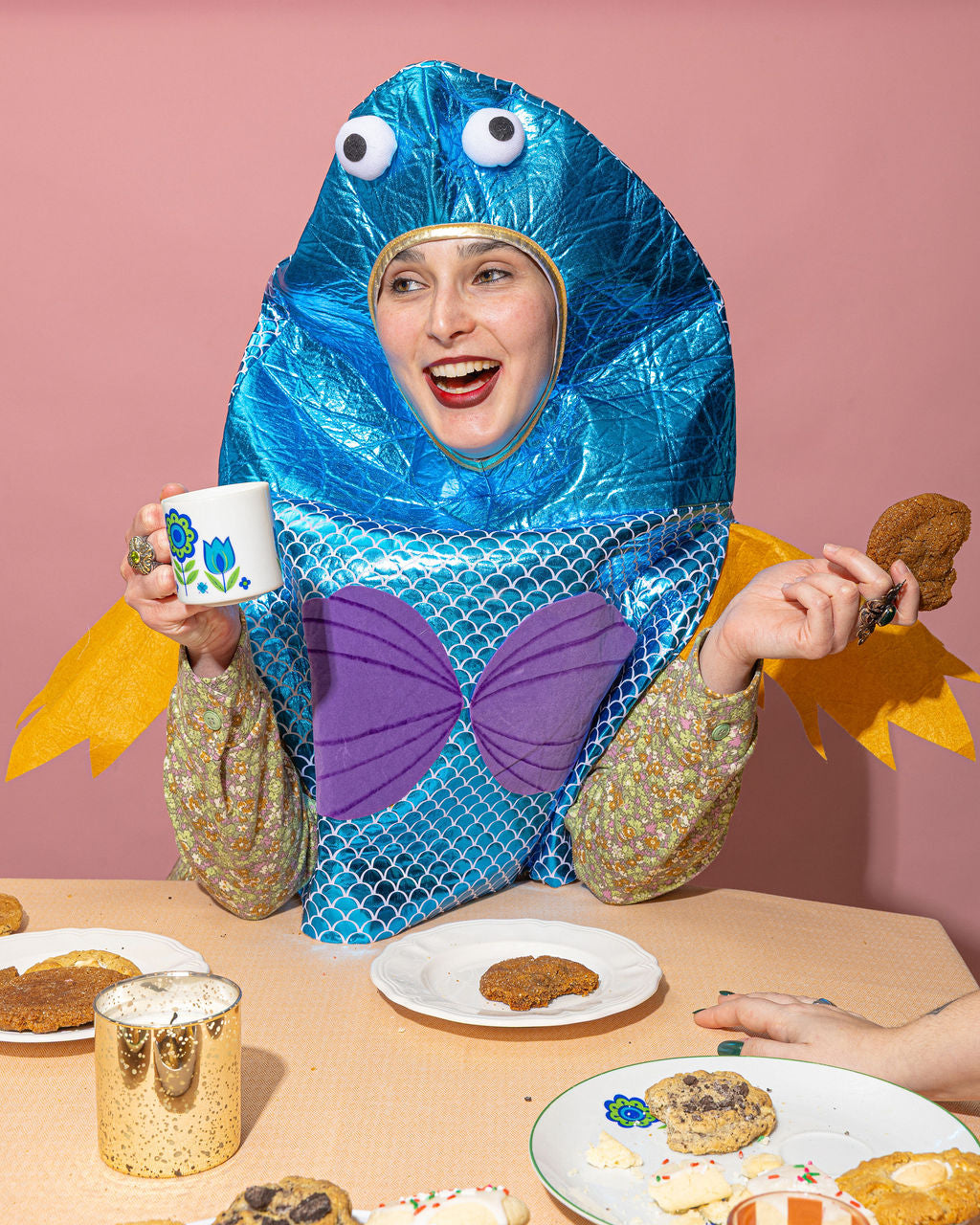 Person in a blue and purple costume with cookie-themed accessories at a table with cookies and a mug.