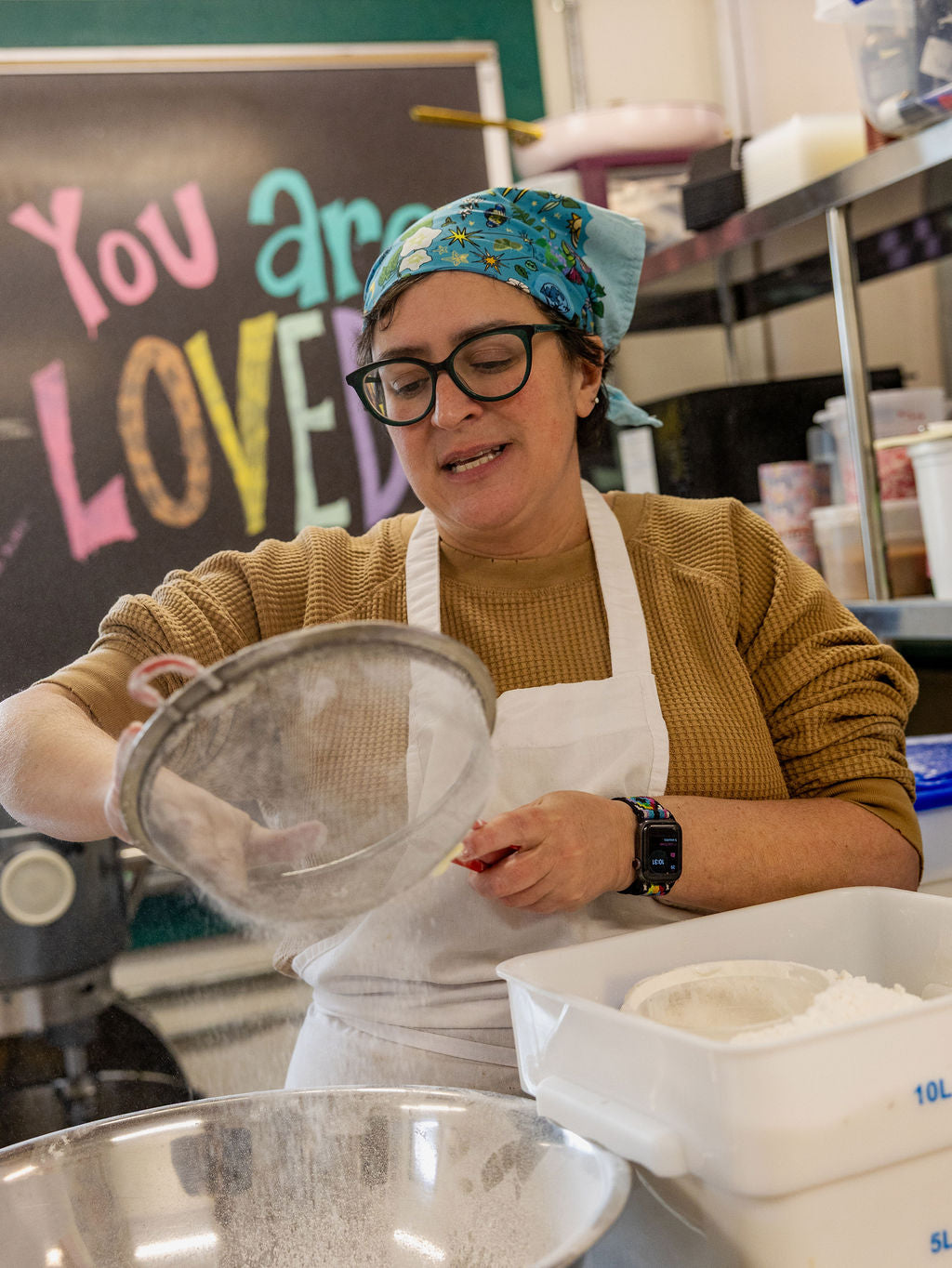 Person in a kitchen wearing an apron and using a strainer, with a sign reading 'You are LOVED' in the background.