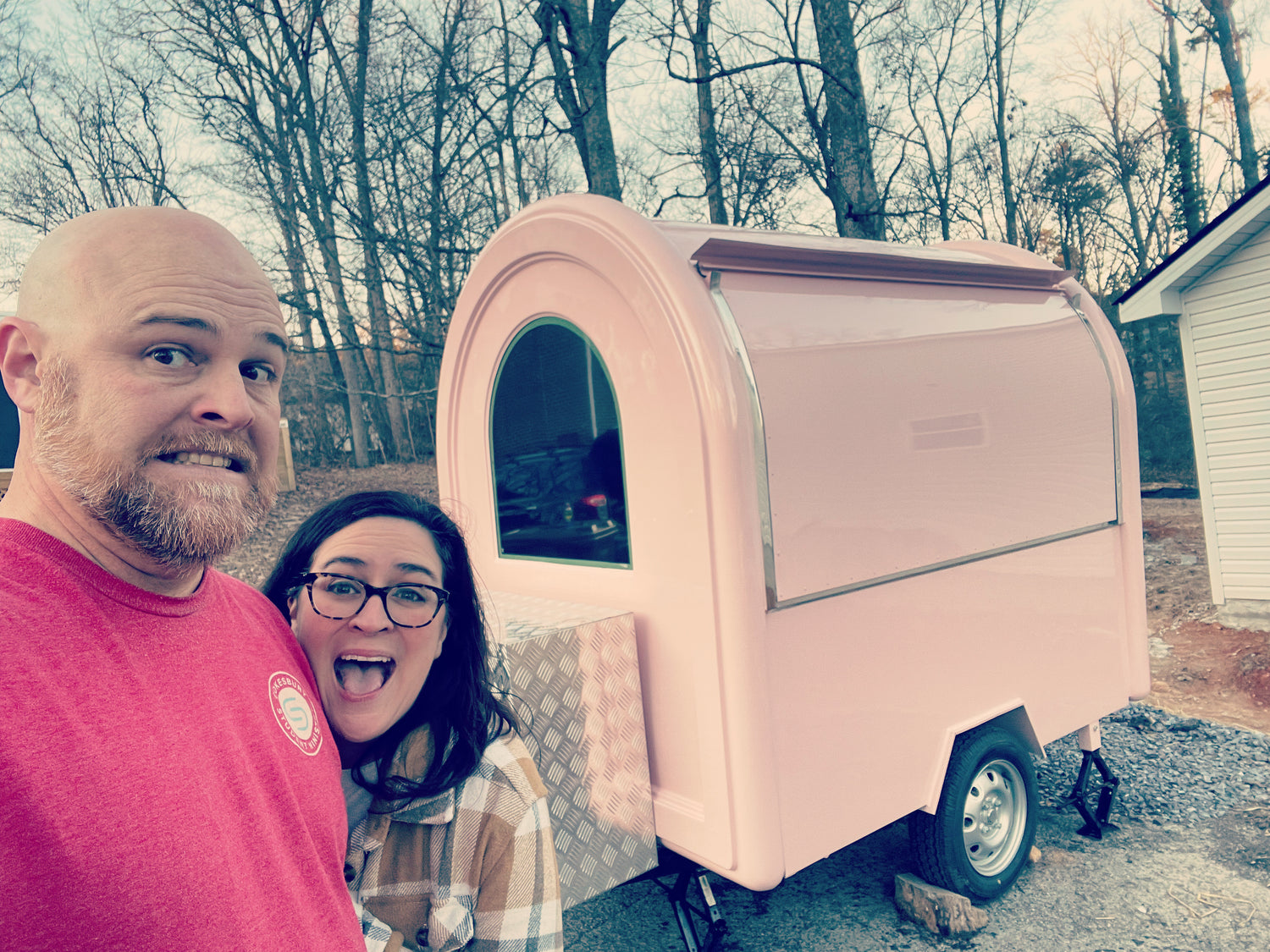 Two people standing in front of a pink trailer that can attach to the back of a vehicle. There is a window that can open on the trailer. 